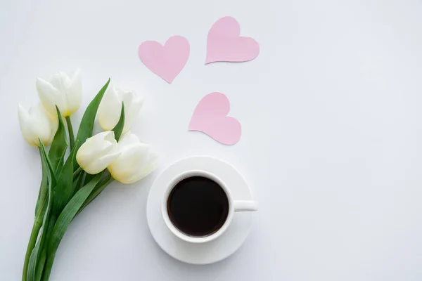 Vista superior de tulipas perto da xícara com café no pires perto de corações de papel no branco — Fotografia de Stock