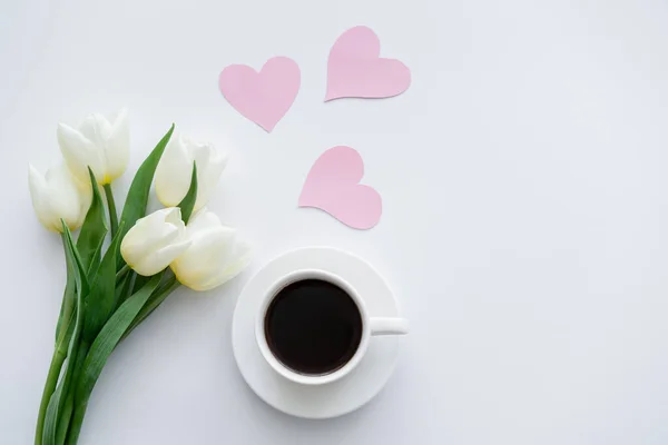 Ansicht von Tulpen in der Nähe von Tasse mit Kaffee auf Untertasse in der Nähe von Papierherzen auf Weiß — Stockfoto