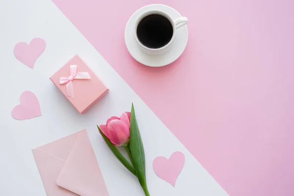 Vue du dessus de l'enveloppe près de tasse avec café, boîte cadeau et tulipe sur blanc et rose — Photo de stock