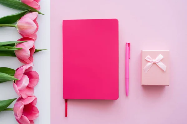 Vista dall'alto della confezione regalo, notebook luminoso e penna vicino ai tulipani su bianco e rosa — Foto stock