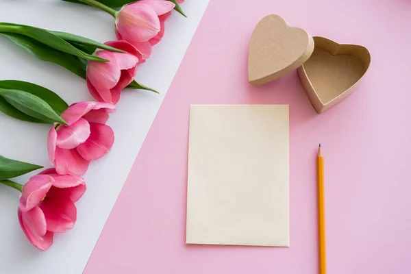 Vista dall'alto di tulipani fioriti vicino scatola regalo a forma di cuore, busta e matita su bianco e rosa — Foto stock