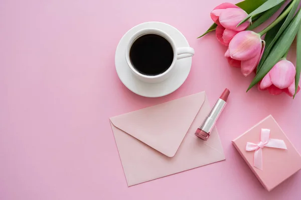 Верхний вид цветущих тюльпанов возле помады, конверт и чашку кофе на розовый — стоковое фото