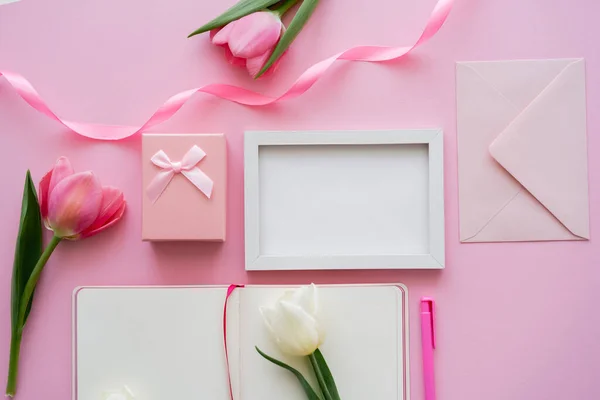 Draufsicht auf Tulpen in Rahmennähe, leeres Notizbuch, Umschlag und Geschenkbox auf rosa — Stockfoto