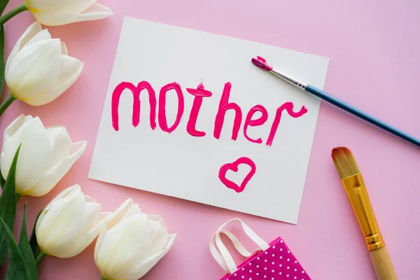 Top view of paintbrush with paint near card with mother lettering and white tulips on pink — Stock Photo