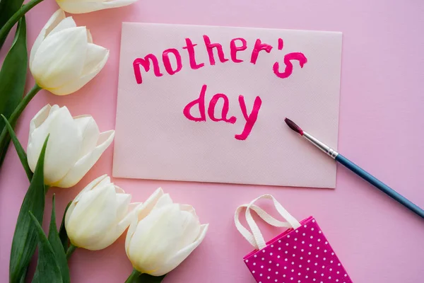 Vista superior do pincel perto do cartão com letras do dia das mães e flores brancas no rosa — Fotografia de Stock
