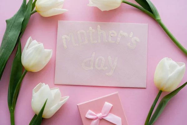 Vista dall'alto della busta con scritte di giorno delle madri vicino alla scatola regalo e fiori bianchi sul rosa — Foto stock