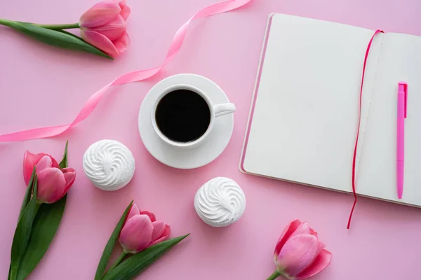 Чашка с кофе рядом с зефиром, пустой блокнот и тюльпаны на розовом — стоковое фото