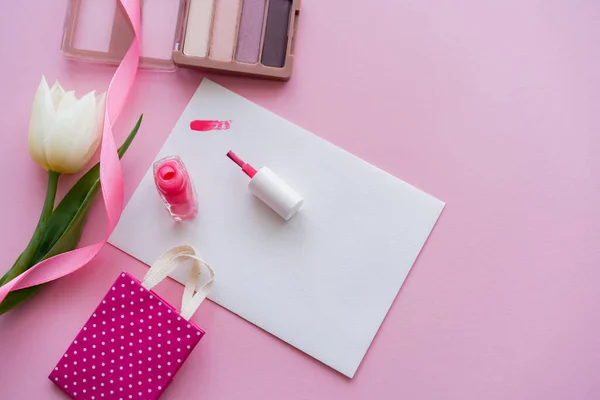 Vue du dessus du coup de pinceau sur papier près du vernis à ongles, tulipe blanche, ombre à paupières et petit sac à provisions sur rose — Photo de stock