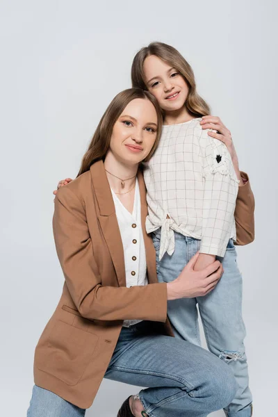 Donna gioiosa e ragazza elegante sorridente alla macchina fotografica mentre abbraccia isolato sul grigio — Foto stock