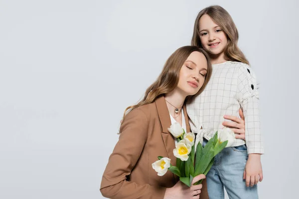 Menina feliz olhando para a câmera perto da mãe com tulipas isoladas em cinza — Fotografia de Stock