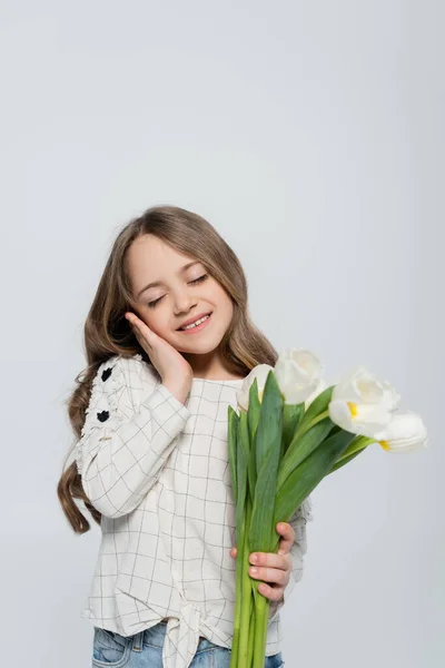 Pleased girl with closed eyes touching face while holding tulips isolated on grey — Stock Photo