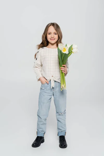 Visão de comprimento total da menina na moda posando com tulipas e mão no bolso no fundo cinza — Fotografia de Stock