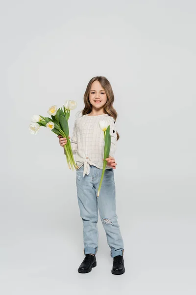 Full length view of girl in jeans and blouse showing white tulip at camera on grey background — Stock Photo