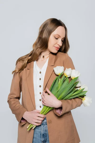 Стильная женщина в коричневом пастельном блейзере позирует с белыми тюльпанами, изолированными на сером — стоковое фото