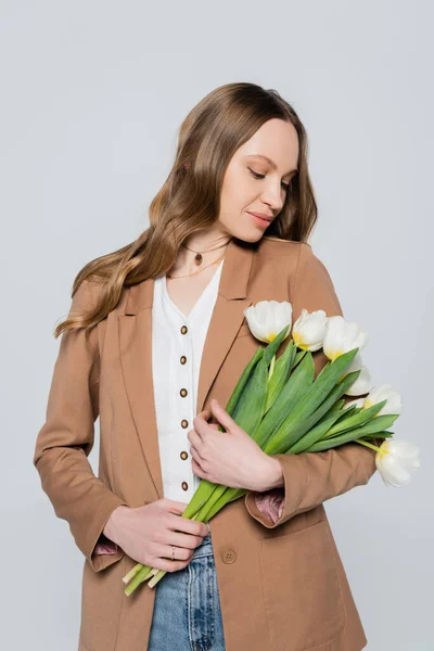 Elegante mujer en blazer marrón pastel posando con tulipanes blancos aislados en gris - foto de stock