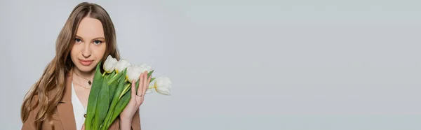 Mulher com cabelos longos olhando para a câmera perto de tulipas frescas isoladas em cinza, banner — Fotografia de Stock