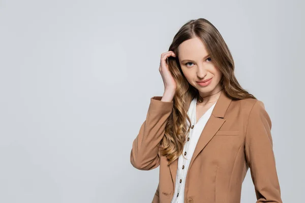 Femme à la mode en blazer brun pastel regardant la caméra et touchant les cheveux longs isolés sur gris — Photo de stock