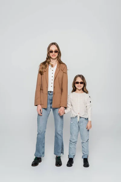 Vue pleine longueur de la mère et de la fille à la mode dans des lunettes de soleil tenant la main sur fond gris — Photo de stock