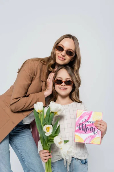 Gioiosa donna in occhiali da sole vicino figlia con tulipani e la giornata delle madri biglietto di auguri isolato su grigio — Foto stock