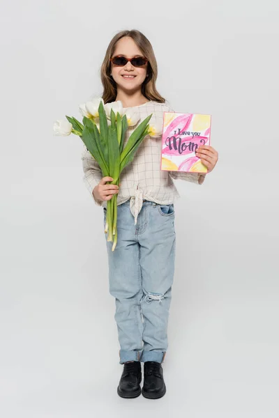 Full length view of happy girl in sunglasses holding tulips and mothers day greeting card on grey background — Stock Photo