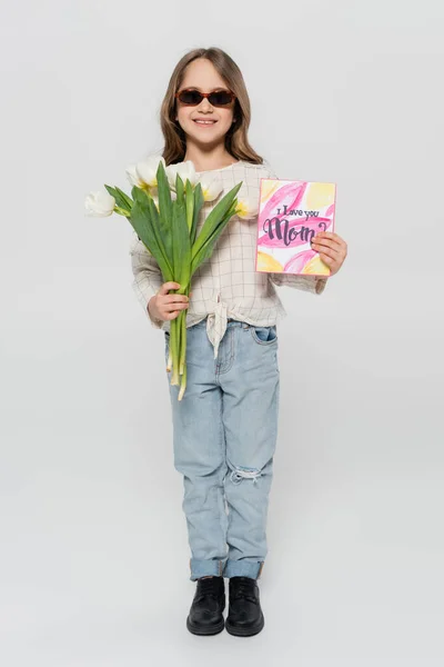 Piena lunghezza vista di ragazza felice in occhiali da sole che tengono tulipani e madri biglietto di auguri giorno su sfondo grigio — Foto stock