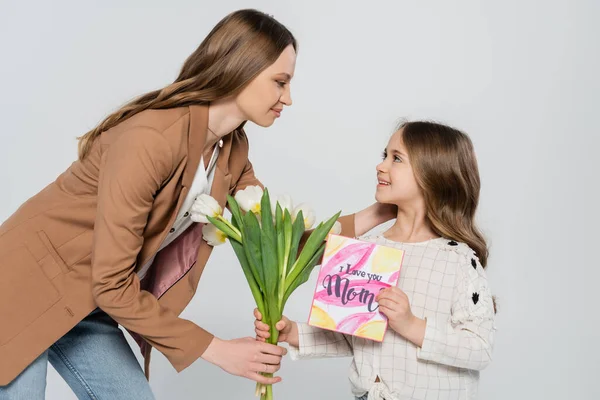 Menina alegre apresentando tulipas e cartão de saudação para a mãe isolado em cinza — Fotografia de Stock