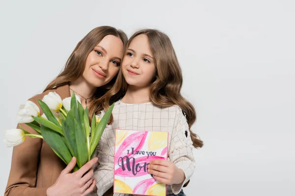 Femme heureuse tenant des tulipes près de sa fille avec carte de vœux de la fête des mères isolé sur gris — Photo de stock