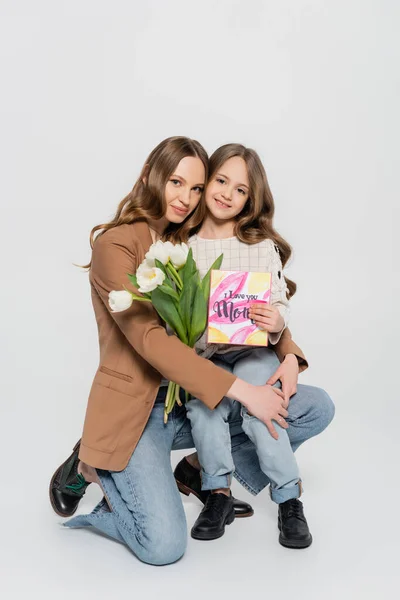 Жінка обіймає дочку, тримає тюльпани та вітальну листівку на сірому фоні — стокове фото