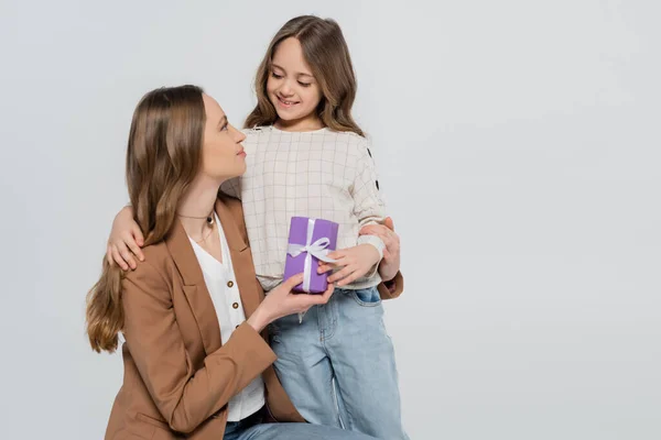 Мать и дочь улыбаются друг другу рядом с подарочной коробкой, изолированной на сером — стоковое фото