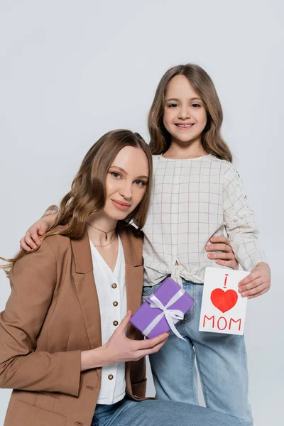 Allegra mamma e figlia con scatola regalo e biglietto di auguri guardando la fotocamera isolata su grigio — Foto stock