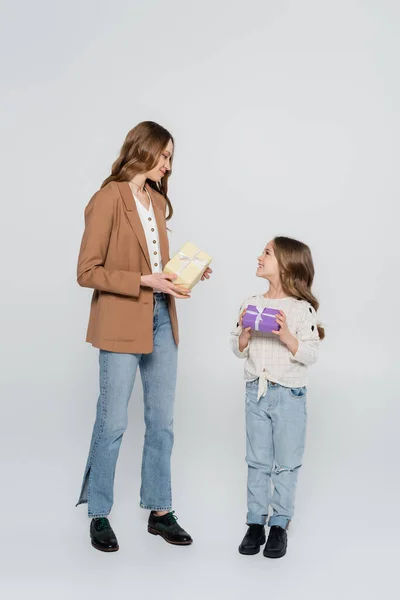 Ganzkörperansicht von stilvoller Mutter und Kind mit Geschenkschachteln, die einander auf grauem Hintergrund anlächeln — Stockfoto