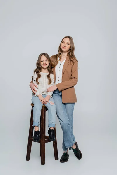 Trendy woman embracing daughter sitting on high stool on grey background — Stock Photo