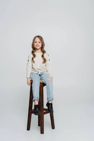 Menina sorridente em blusa branca e jeans sentado em fezes altas em fundo cinza — Fotografia de Stock