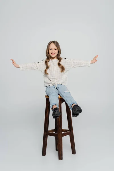 Visão de comprimento total da menina alegre sentada em alto banco com as mãos estendidas no fundo cinza — Fotografia de Stock