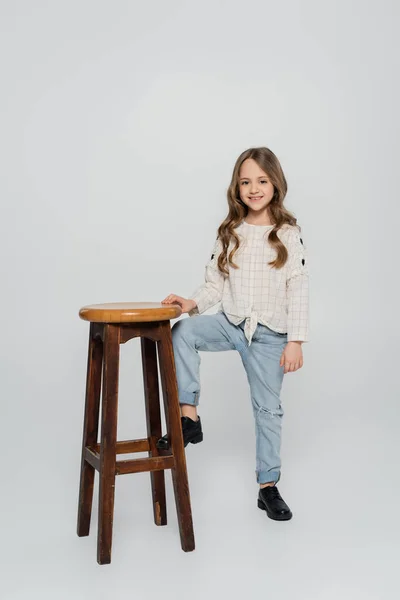 Полный вид стильного ребенка, позирующего рядом с высоким стулом на сером фоне — стоковое фото