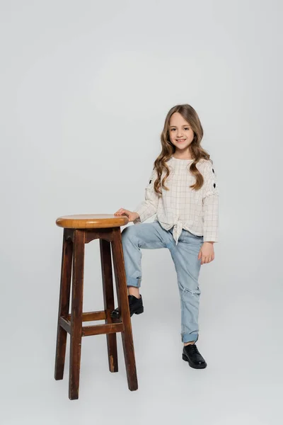 Vue pleine longueur de l'enfant élégant posant près de haut tabouret sur fond gris — Photo de stock