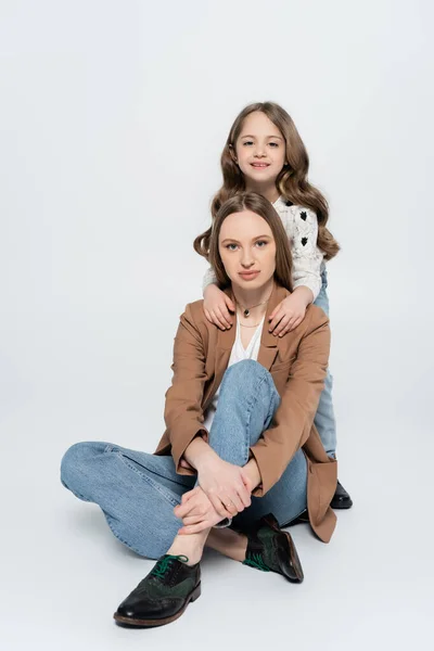 Mädchen umarmt Schultern der Mutter sitzend mit überkreuzten Beinen auf grauem Hintergrund — Stockfoto