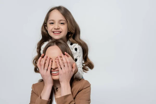 Fröhliches Mädchen, das die Augen einer lächelnden Mutter verdeckt, während es isoliert auf grau in die Kamera blickt — Stockfoto