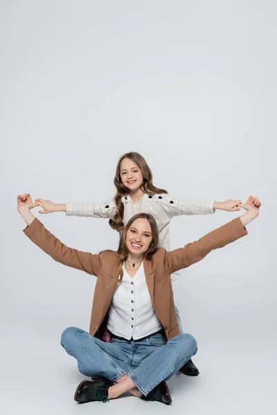 Aufgeregtes Mädchen hält Hände mit glücklicher Mutter, die mit überkreuzten Beinen auf grauem Hintergrund sitzt — Stockfoto