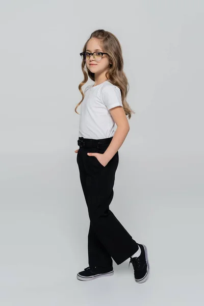 Полный вид девушки в очках, позирующей с руками в карманах черных брюк, изолированных на сером — стоковое фото