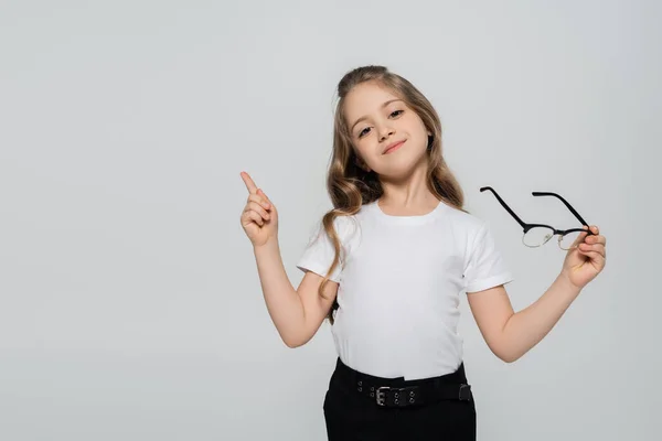 Smiling child holding eyeglasses and pointing with finger isolated on grey — Stock Photo