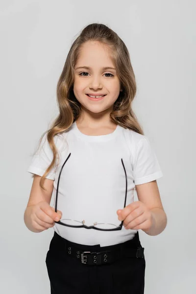 Cheerful girl in white t-shirt holding eyeglasses and looking at camera isolated on grey — Stock Photo