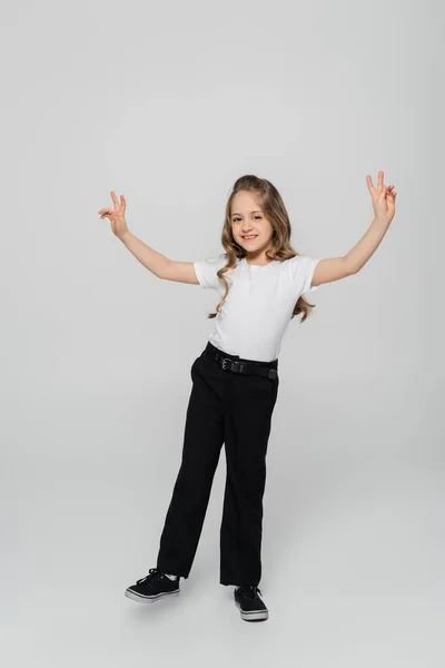 Vue pleine longueur de fille heureuse en pantalon noir montrant des signes de victoire sur fond gris — Photo de stock