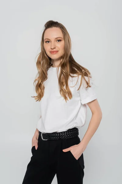 Pretty woman with long hair holding hands in pockets of black trousers isolated on grey — Stock Photo