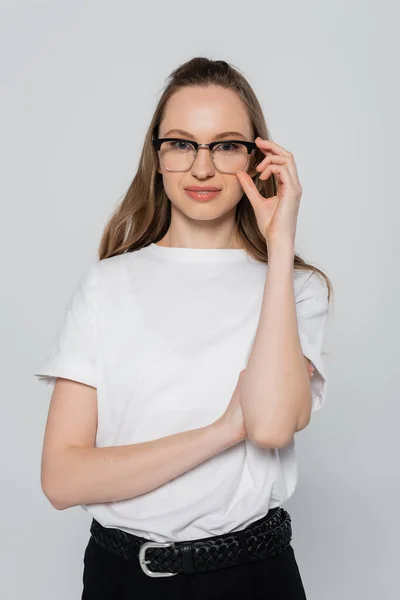 Mulher positiva em t-shirt branca ajustando óculos isolados em cinza — Fotografia de Stock