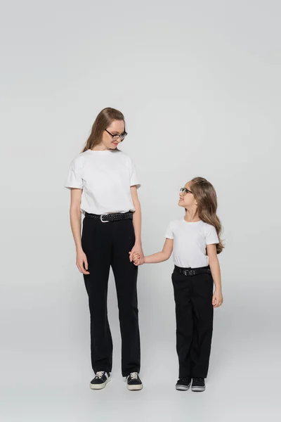 Visão de comprimento total de mãe e filho de mãos dadas e olhando uns para os outros em fundo cinza — Fotografia de Stock