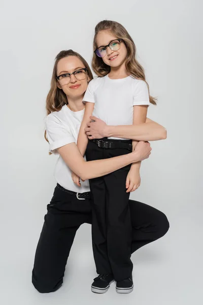 Mamá y su hija en gafas graduadas y ropa en blanco y negro mirando a la cámara sobre fondo gris - foto de stock