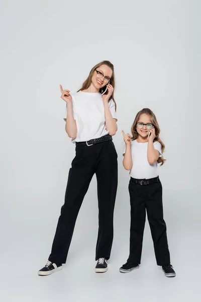 Ganzkörperansicht von Mutter und Tochter, die auf Mobiltelefonen sprechen und mit den Fingern auf grauem Hintergrund zeigen — Stockfoto