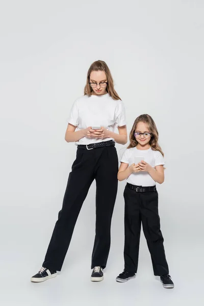 Vista completa de la madre y la hija en pantalones negros charlando en teléfonos inteligentes sobre fondo gris - foto de stock