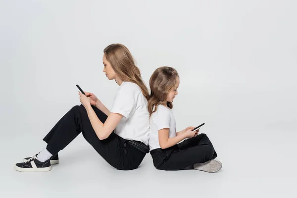 Side view of mother and child using smartphones while sitting back to back on grey background — Stock Photo